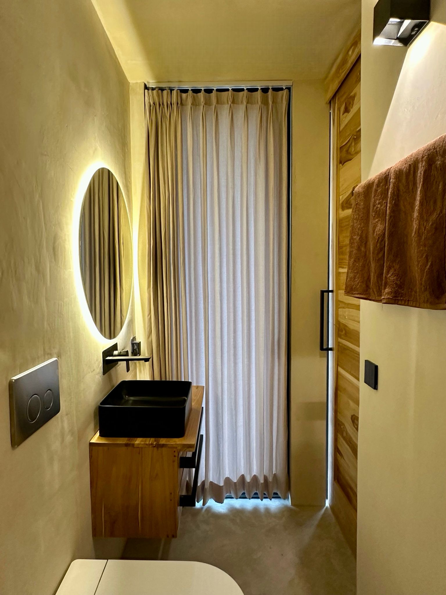 Warm-toned bathroom with microcement walls, round backlit mirror, wood-clad door, black sink on wood vanity