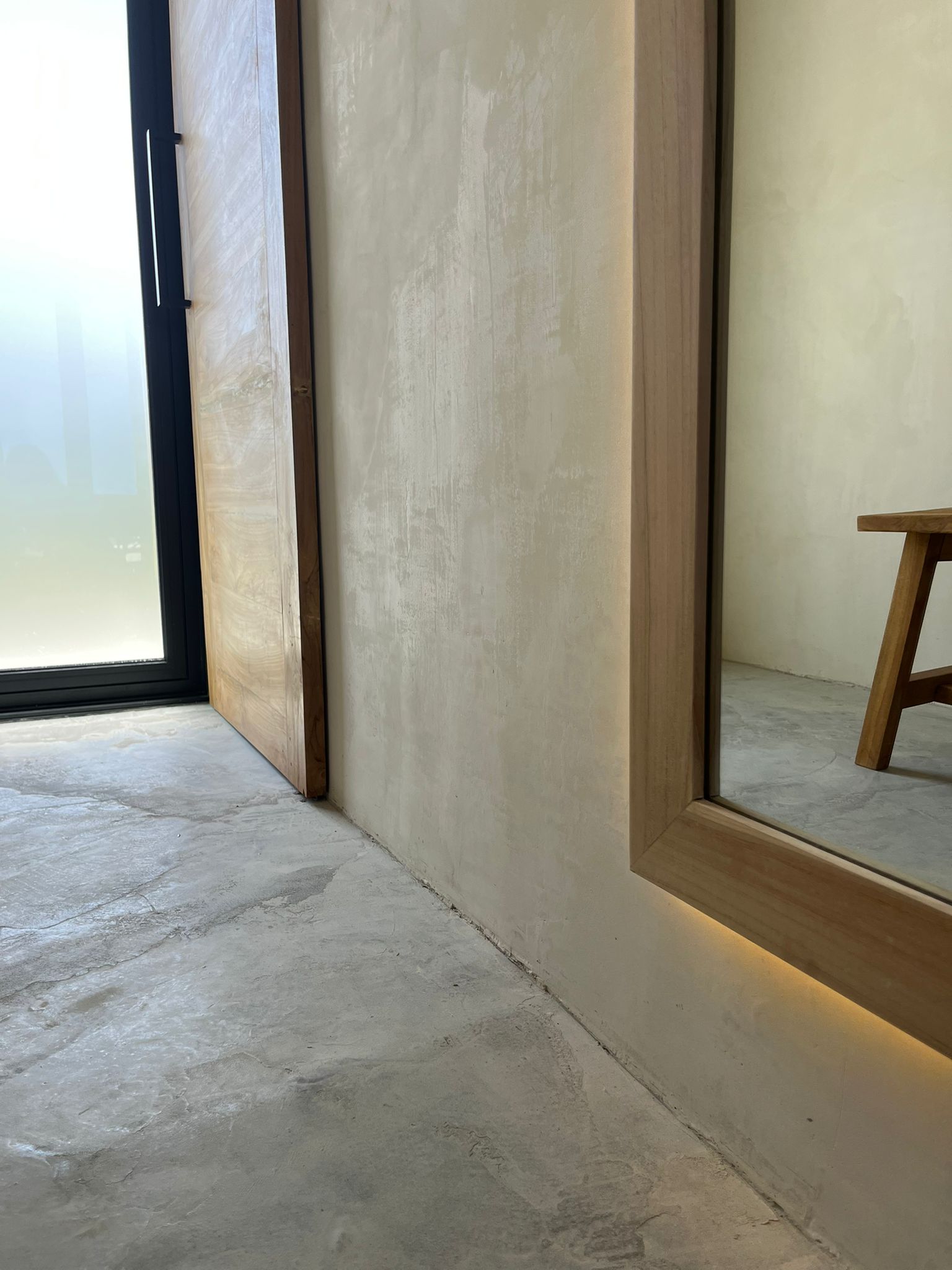 Warm beige microcement floor and wall meeting a wood-framed mirror with hidden uplighting